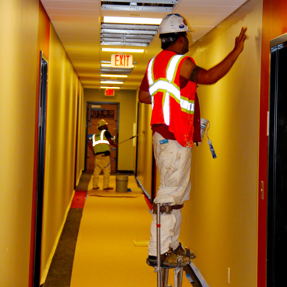 Maintenance & Construction. A pair of painters, one on stilts and the other with a long pole, roll paint onto the walls of a hallway.